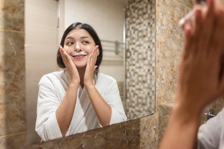 Indonesian southeast asian woman wrapped in a towel, looking at herself in the mirror while applying some cream. Natural skincare and wellness conceptの写真素材