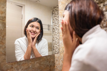 Indonesian southeast asian woman wrapped in a towel, looking at herself in the mirror after washing and cleaning her face with splashing water. Natural skincare and wellness conceptの写真素材