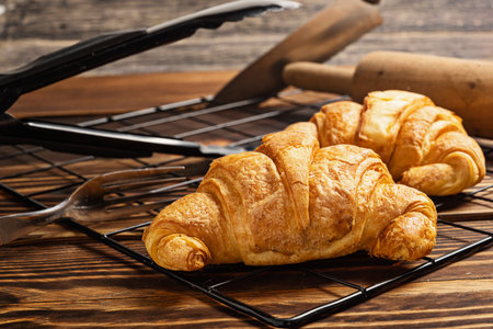 Freshly baked croissants on a cooling tray with tongs and a rolling pin on the tableの写真素材