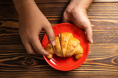 Human hand holding a freshly baked croissant on a red plate with a wooden background. Top viewの写真素材