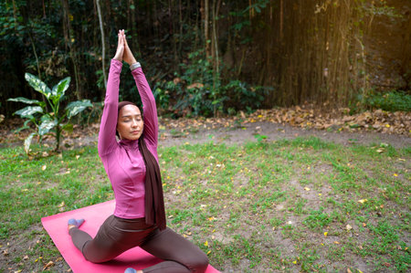 Indonesian southeast asian muslim woman in a sporty outfit practices yoga on a fitness mat in a public park outdoors. Concept of a Healthy active lifestyleの写真素材