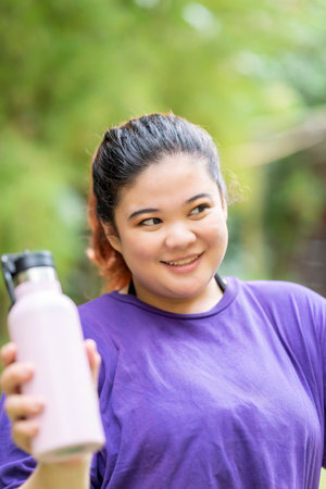 Indonesian southeast asian overweight woman in a sporty outfit drinking water after practice running exercise in a public park outdoors. Concept of a Healthy active lifestyle and body inclusivityの写真素材