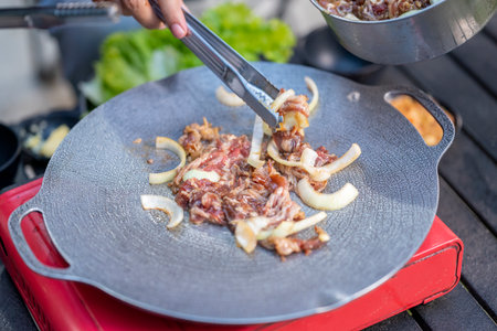 Closeup view of people with tongs cooking sliced marinated beef bulgogi on a round grill pan with onions. The scene of the lifestyle of enjoying food in a natural settingの写真素材