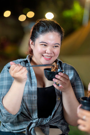 Indonesian southeast asian woman enjoying a meal, using chopsticks to serve themselves a barbecue meal from small black bowls. The scene of the lifestyle of enjoying food in a natural settingの写真素材