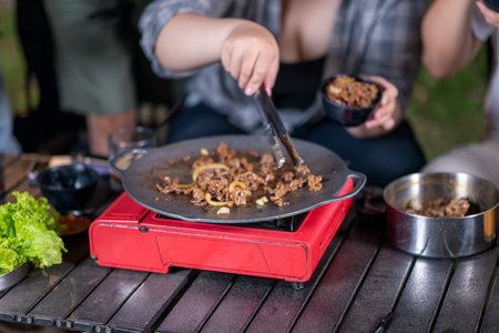 Closeup view of people with tongs cooking sliced marinated beef bulgogi on a round grill pan with onions. The scene of the lifestyle of enjoying food in a natural settingの写真素材