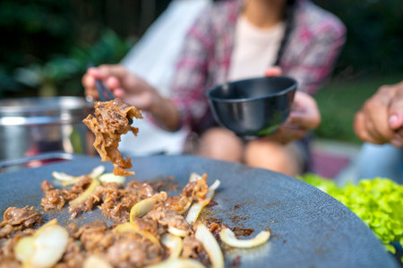 Closeup view of people with chopsticks cooking sliced marinated beef bulgogi on a round grill pan with onions. The scene of the lifestyle of enjoying food in a natural settingの写真素材