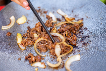 Closeup view of people with chopsticks cooking sliced marinated beef bulgogi on a round grill pan with onions. The scene of the lifestyle of enjoying food in a natural settingの写真素材