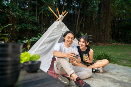 Indonesian southeast asian couple smiles while sitting together outdoors and enjoying refreshing drinks. A man holds the cellphone. The scene of the lifestyle of enjoying food in a natural settingの写真素材