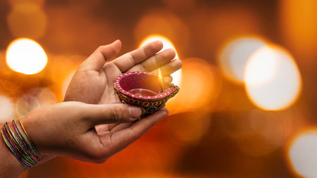 Woman hand holding a lit clay diya lamp with a blurred bokeh light background. Celebration of Diwali...