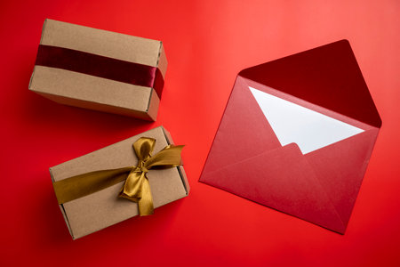 Top view of brown gift box tied with golden and red ribbon with red envelope on colored background....