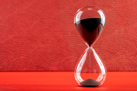 Hourglass with black sand on a red background. Time passing or time flowing. Sand glass, sand timer,...