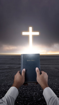 Prayer holding the blue book of the holy bible with light of the Christian cross on dark dramatic sky background. Concept of Christianity, religious, faith, Jesus, or beliefの写真素材