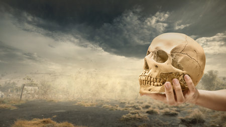 Human hand holding human head skull with dark and dramatic cloudscape background. The scene is scary and mysterious. Concept of Spooky Halloweenの写真素材