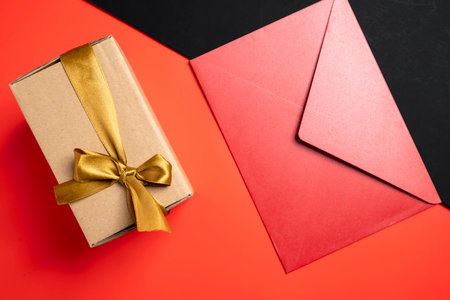Top view of a brown gift box tied with a golden ribbon and a red envelope on a colored background....