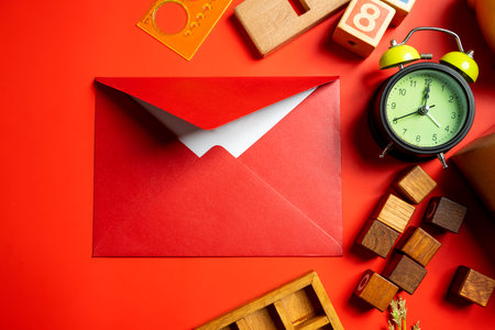 Top view of red envelope or postcard with green alarm clock and wooden cubes on red background. Mockup, blank template, copy space. Greeting or invitation card with flat lay styleの写真素材