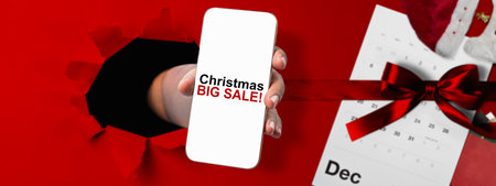 Human hand through hole paper showing Christmas big sale text on cellphone screen with red ribbon. Copy space. Christmas sale conceptの写真素材