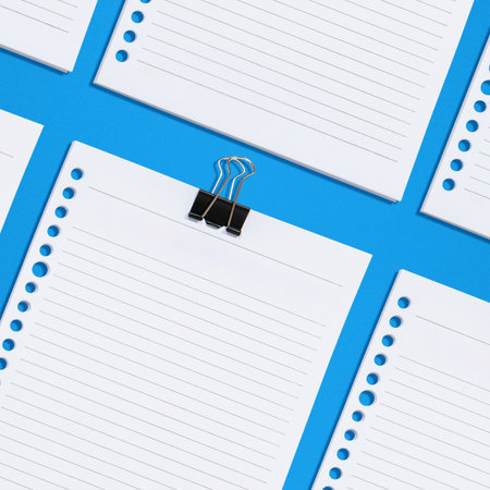 Flat lay composition featuring several lined paper sheets with hole punches, one held together with a black binder clip, against a bright blue background. Mockup or Copy spaceの写真素材