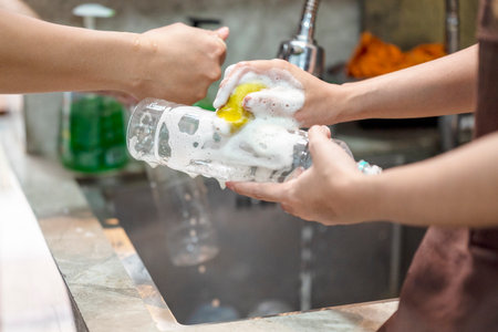 Person hands washing a clear plastic bottle with a yellow sponge under a stream of water in a kitchen sink, promoting cleanliness and hygieneの写真素材