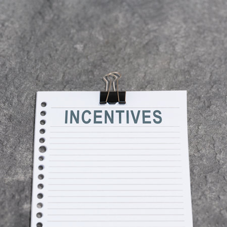 Top view image of a notepad sheet with the word "Incentives" printed on it, attached with a binder clip, placed on a textured concrete surfaceの写真素材