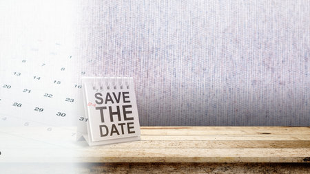 A white spiral desk calendar with 'Save The Date' text stands on a wooden table against a textured wall background, offering plenty of copy space for event announcements and remindersの写真素材