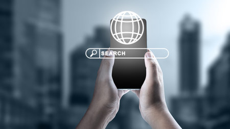Hands holding a mobile phone with a digital search bar and globe icon against a blurred urban background. Concept of global information technology, connectivity, and internet searchの写真素材
