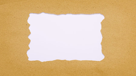 A piece of textured brown kraft paper with a torn rectangular hole in the center, revealing white background. Ideal for copy space, templates, and creative designsの写真素材