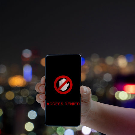 Hand holding a smartphone displaying an Access Denied message and stop icon against a blurred night...