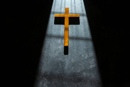 Simple wooden Christian cross on a dark textured surface, dramatically illuminated by a bright shaft of light from a doorway, symbolizing faith, hope, and spiritual light in the darknessの写真素材