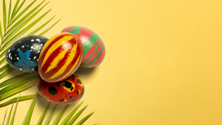 Creative flat lay of four hand-painted Easter eggs and green leaves on a vibrant yellow background....