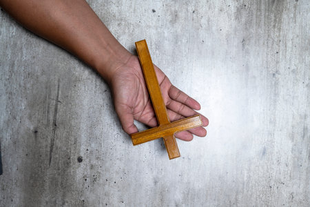 A dark-skinned hand holds a small wooden inverted cross against a weathered gray concrete floor,...