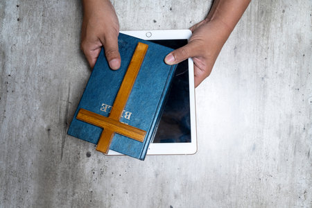 Top down view of hands holding a blue Holy Bible and wooden cross over a white tablet on a grey...