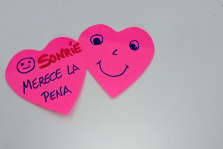 Two pink heart-shaped sticky notes with hand-drawn cheerful faces and the Spanish text "SonrÃ­e, Merece la Pena" (Smile, It's Worth It) encourage positivity.の写真素材