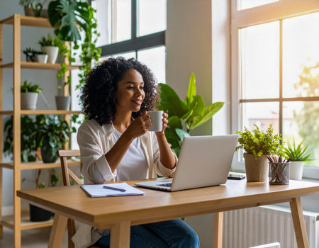 Smiling businesswoman drinking coffee while working on laptop in home office full of plants, enjoying break and looking outside windowの素材