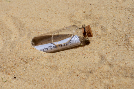 Glass bottle with cork stopper containing rolled-up paper with geographic coordinates, lying on beach sandの写真素材