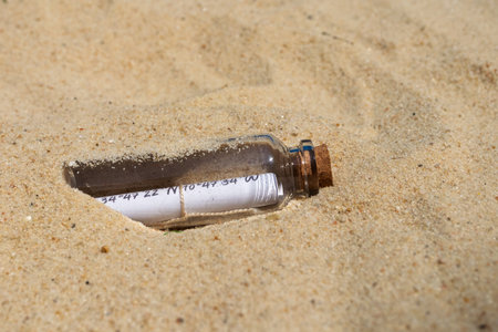 Glass bottle with cork lid containing rolled-up paper with handwritten coordinates partially buried in beach sandの写真素材