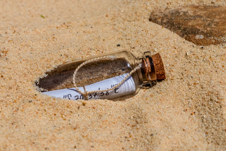 Glass bottle with cork stopper containing rolled-up paper with handwritten coordinates, half-buried in beach sandの写真素材