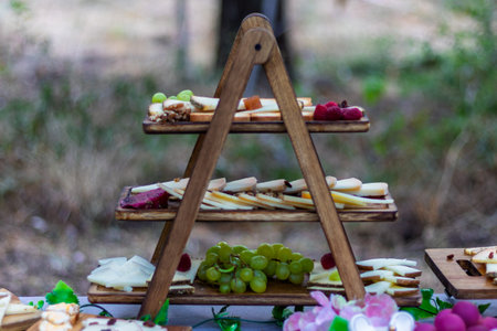 Outdoor catering setup displaying a rustic three-tier stand filled with various cheeses, fresh grapes, and berries for guestsの写真素材