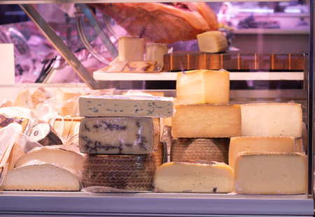 Artisanal cheese selection stacked and unwrapped, presented in a refrigerated display case at a traditional food marketの写真素材