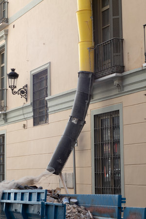 Building renovation creating rubble, with a debris chute directing discarded materials into a large container on the streetの写真素材