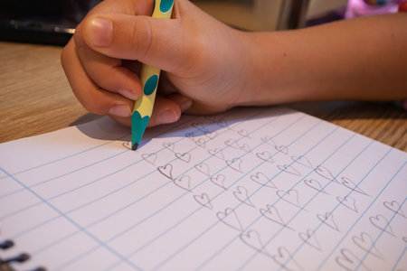 Child hand holding a pencil, repetitively drawing heart shapes on a notebook page, symbolizing love and learningの写真素材