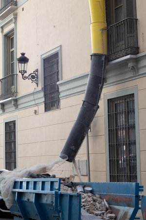 Building renovation site with a debris chute directing demolition waste into a large blue skip container on an urban streetの写真素材