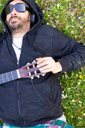 Hippy man lying down with beard and glasses playing guitar outdoors on the grass