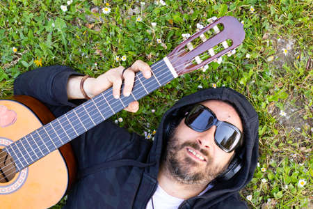 Hippy man lying down with beard and glasses playing guitar outdoors on the grass
