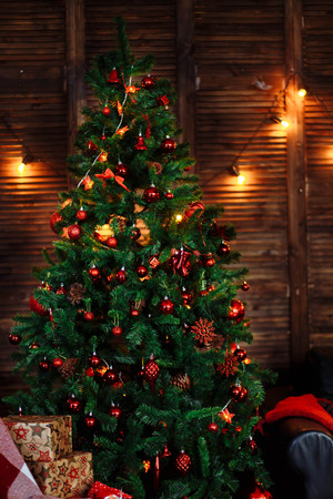 green Christmas tree decorated with red toys, ornaments, pine cones, beads garlands box sofa wooden wall floor in the darkの写真素材