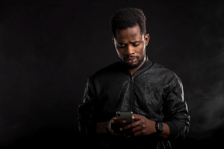 Portrait of good-looking happy african american in jacket, holding phone and gazing at screen, sending text message from friend, reading good news against black background. Spare time and technology.の写真素材