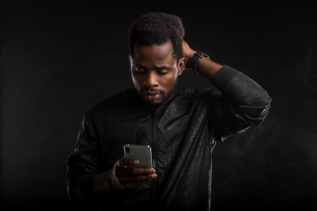 Indoor shot of impressed and amazed attractive African American man with beard and short haircut, holding smartphone, looking surprised at gadget screen with open mouth in dark. Emotions, technology.の写真素材