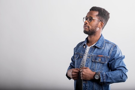 Side view portrait of serious confident unshaven african man with full lips, short curly hair wearing eyewear, blue stylish denim jacket, watch, looking away on gray background with copy space.の写真素材