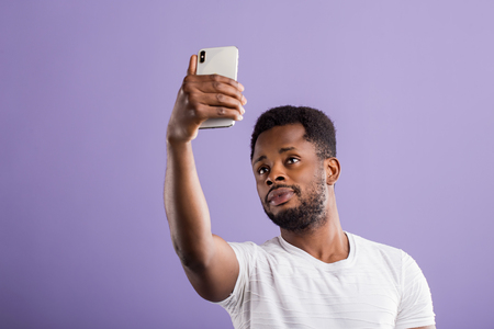 Portrait of confident attractive friendly african american student with short curly hair and beard wearing white t shirt holding mobile phone taking selfie. Technology and communication concept.の写真素材