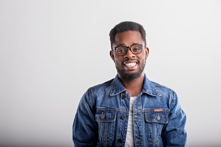 Horizontal close up portrait of joyful happy african guy in spectacles denim jacket smiling broadly with teeth laughing looking at camera over gray background. Facial expressions and emotions concept.の写真素材