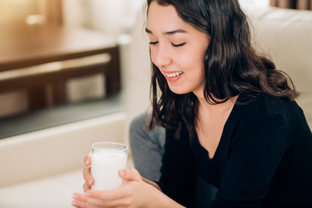 Joifull satisfied female in casual clothes enjoying morning weekend. Close up shot of beautiful student with glass of white smoothie. Woman take care of her health. Diet, vitamins, healthy drink.の写真素材